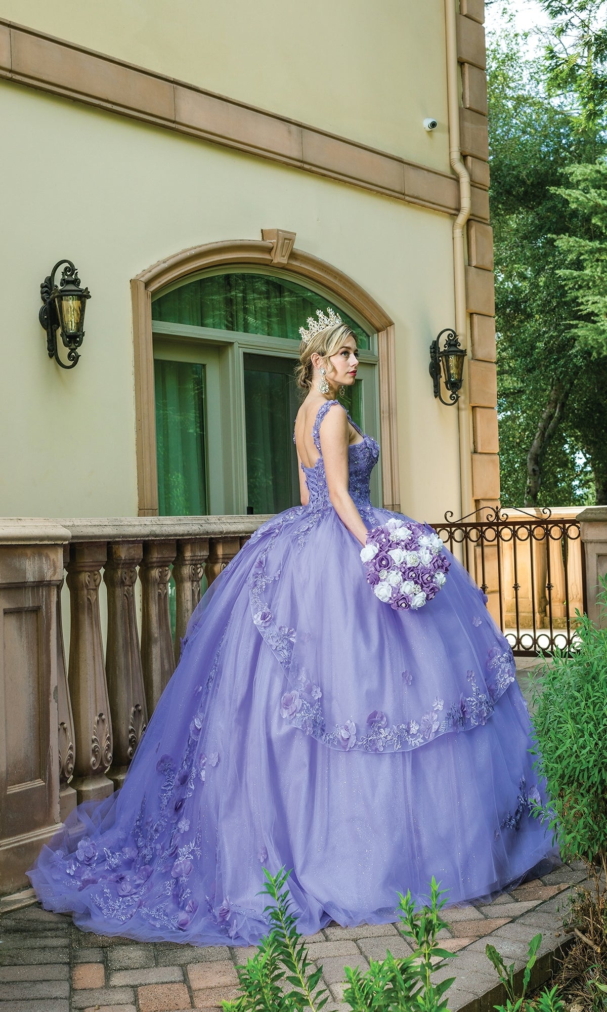A woman in the Quinceanera Dress 1627 By Dancing Queen and a tiara stands on a stone patio, holding a bouquet of purple and white flowers, with floral details and greenery adding elegance to the scene.